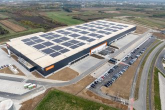 Pogled iz zraka na veliku sivu skladišnu zgradu sa solarnim panelima na krovu, utovarnim rampama i velikim parkiralištem.