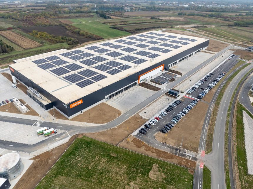 Pogled iz zraka na veliku sivu skladišnu zgradu sa solarnim panelima na krovu, utovarnim rampama i velikim parkiralištem.