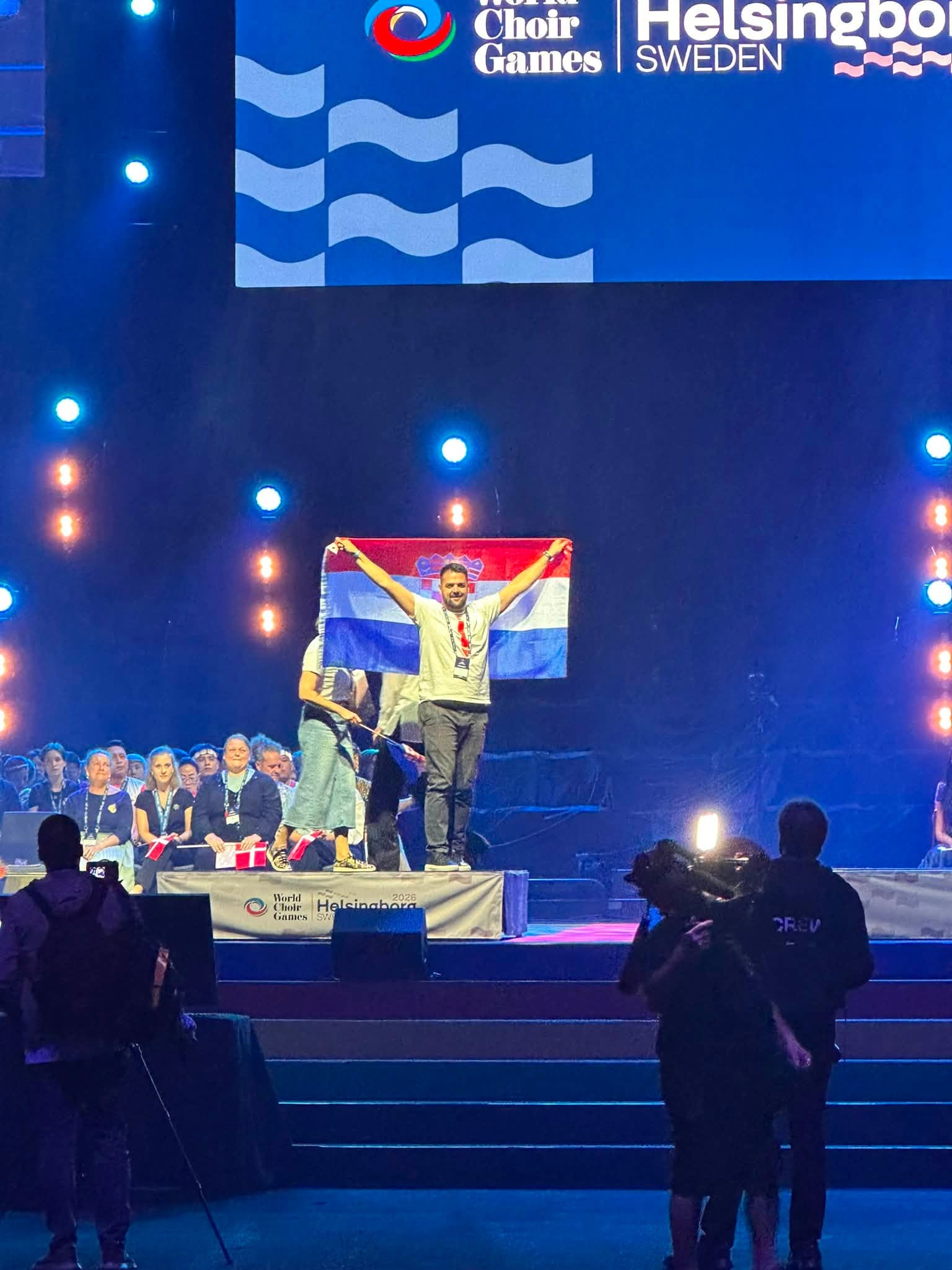 Muškarac na osvijetljenoj pozornici visoko drži hrvatsku zastavu ispred velikog ekrana s natpisom World Choir Games Helsingborg Sweden.