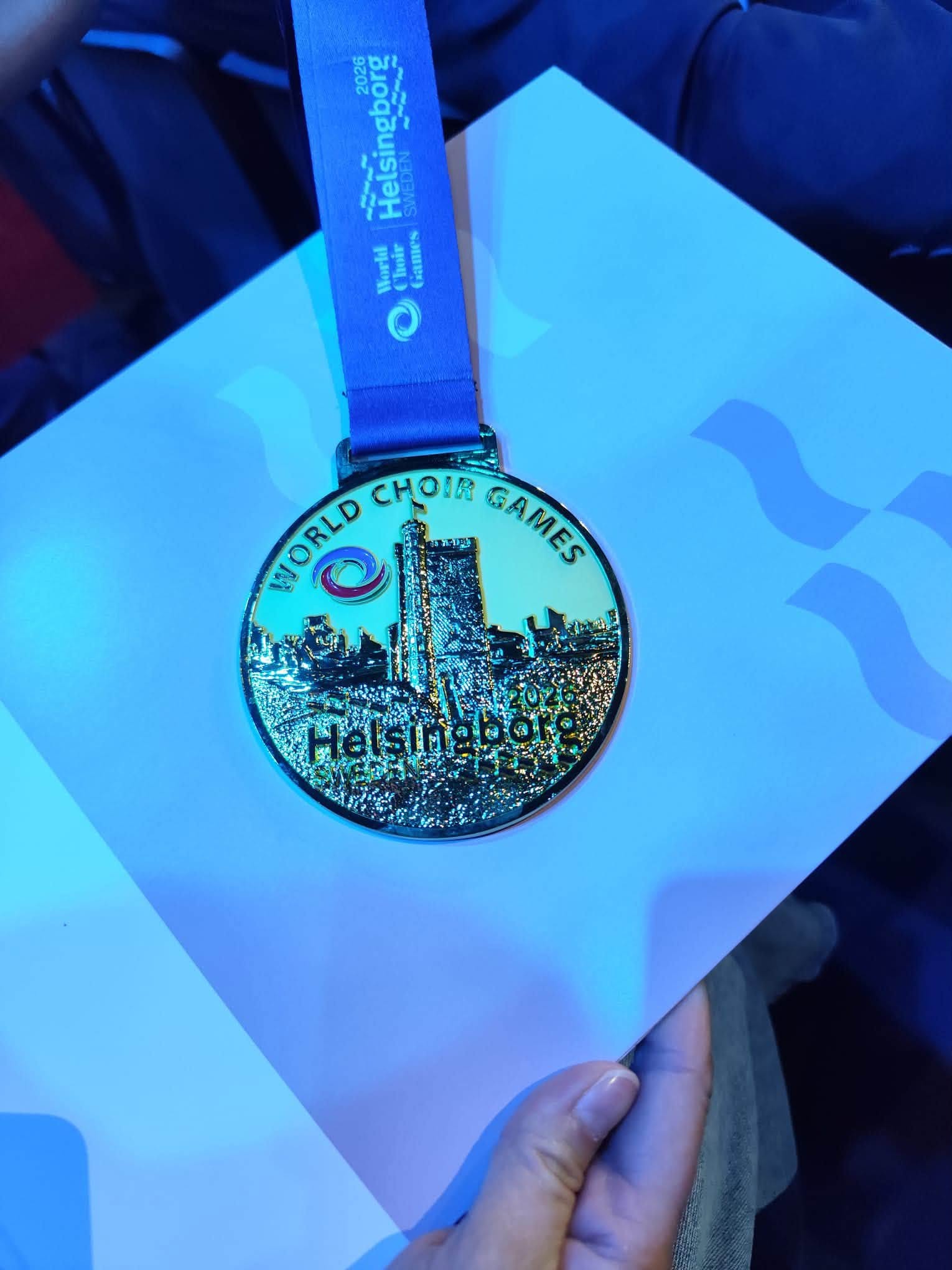 Zlatna medalja s plavom trakom na kojoj piše World Choir Games Helsingborg 2026 Švedska, u ruci s plavim osvjetljenjem.