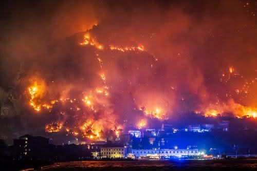 Pogled noću na vatrenu stihiju i dim na brdu iznad kuća i obale uz prisutnost hitnih službi.