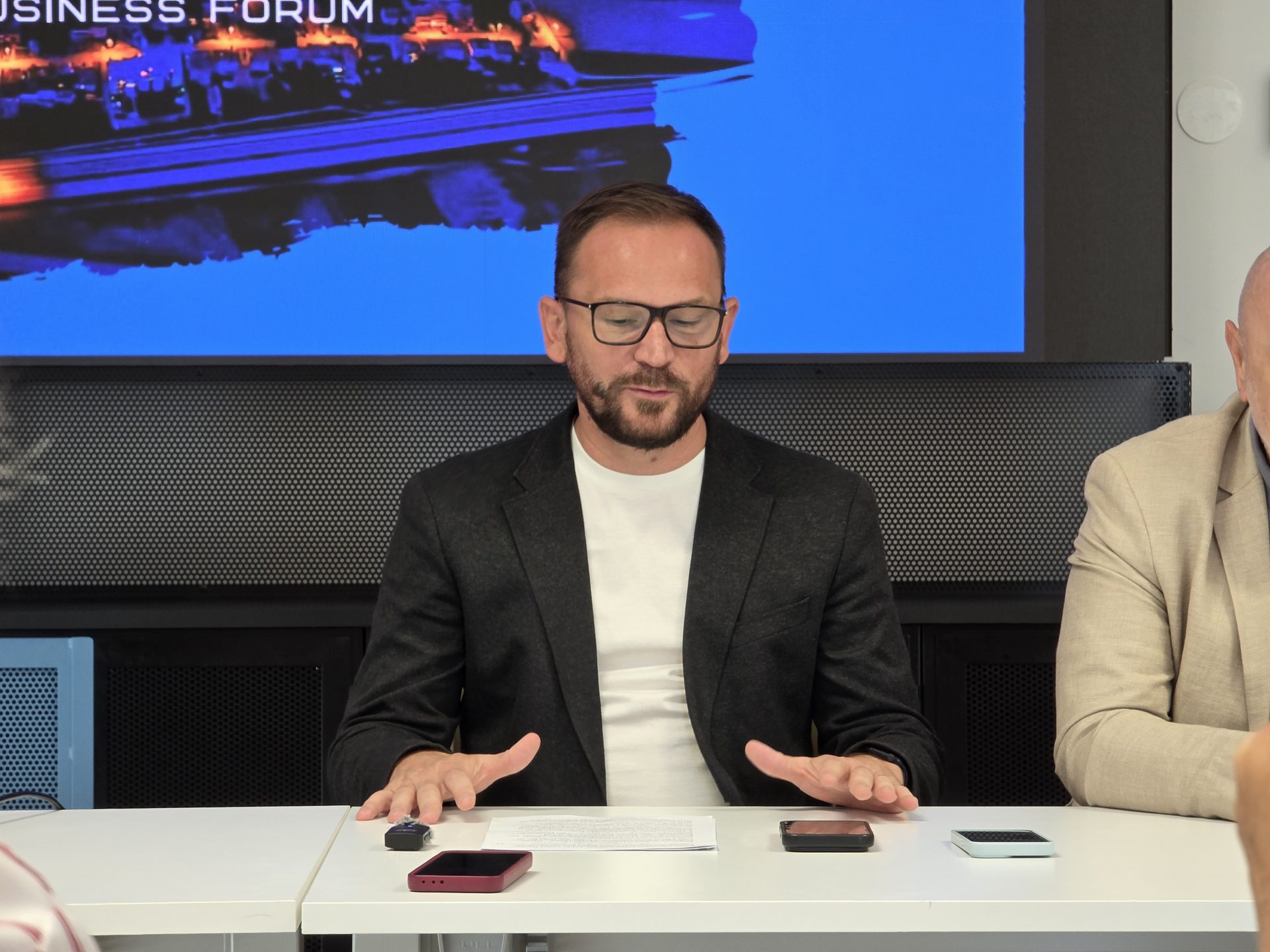 Muškarac u crnom sakou s naočalama gestikulira rukama sjedeći za konferencijskim stolom ispred plavog ekrana.