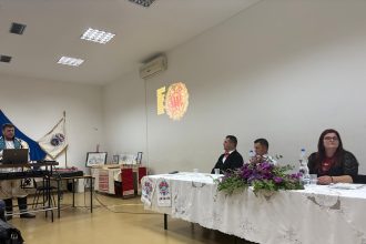 Muškarac u narodnoj nošnji stoji uz laptop i prezentira, dok troje ljudi sjedi za ukrašenim stolom u dvorani.