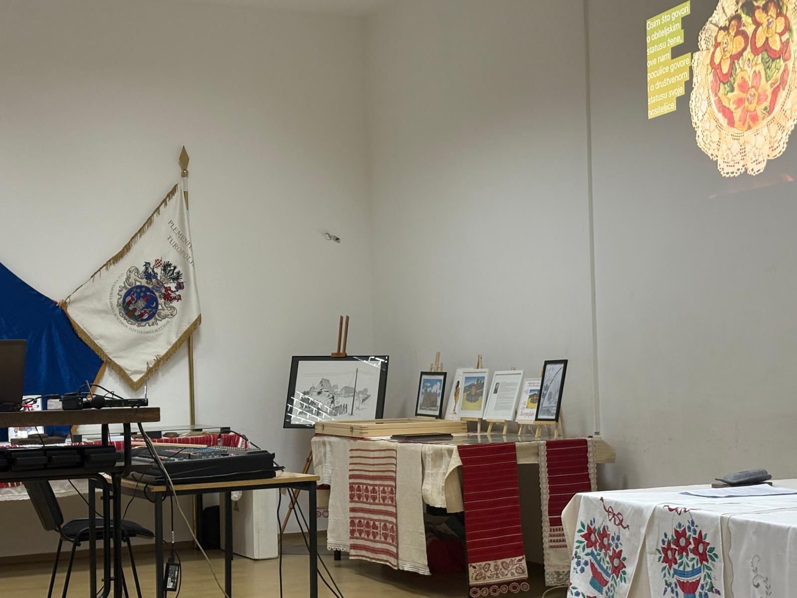 Prostorija pripremljena za kulturno predavanje s izloženim uokvirenim slikama na vezenim stolnjacima, zastavom Plemenite opčine Turopolje i projekcijom o poculici na zidu.