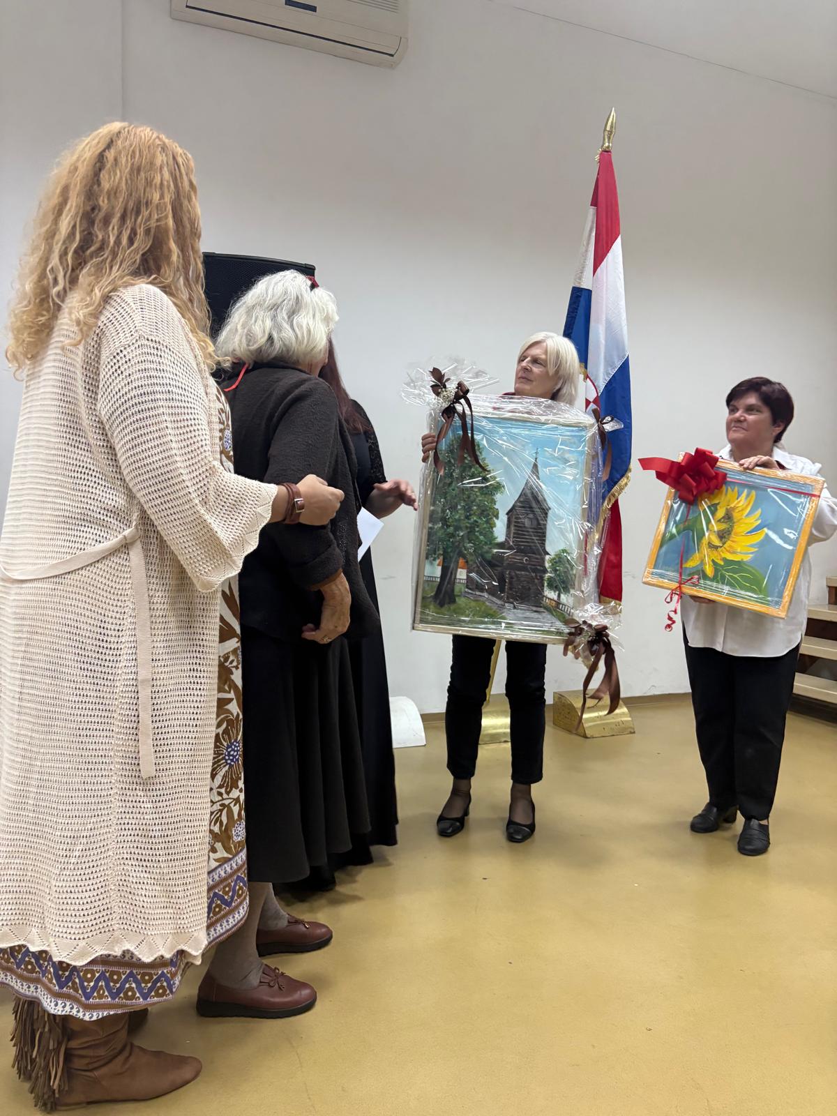 Grupa žena u dvorani, od kojih dvije u rukama drže uokvirene umjetničke slike umotane u celofan s mašnama, dok u pozadini stoji zastava Republike Hrvatske.