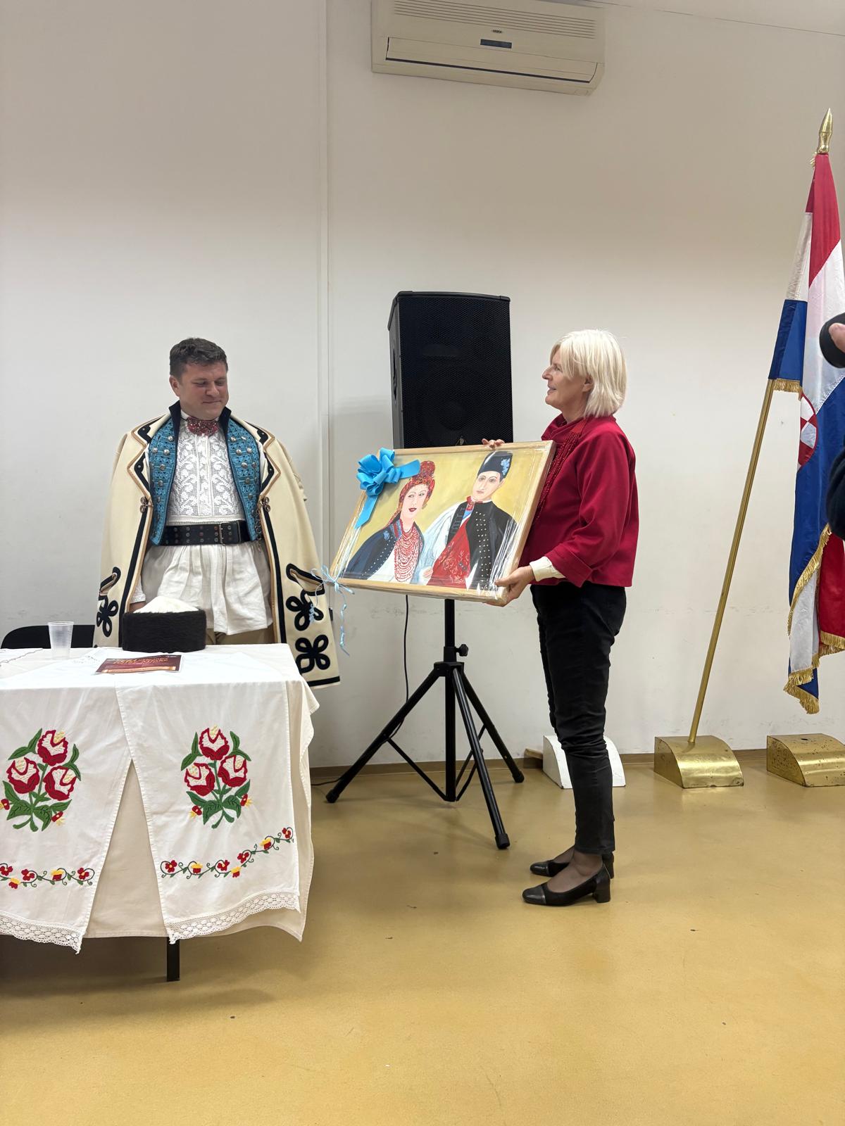 Žena drži uokvirenu sliku s motivom para u narodnoj nošnji pored muškarca odjevenog u tradicijsku nošnju.